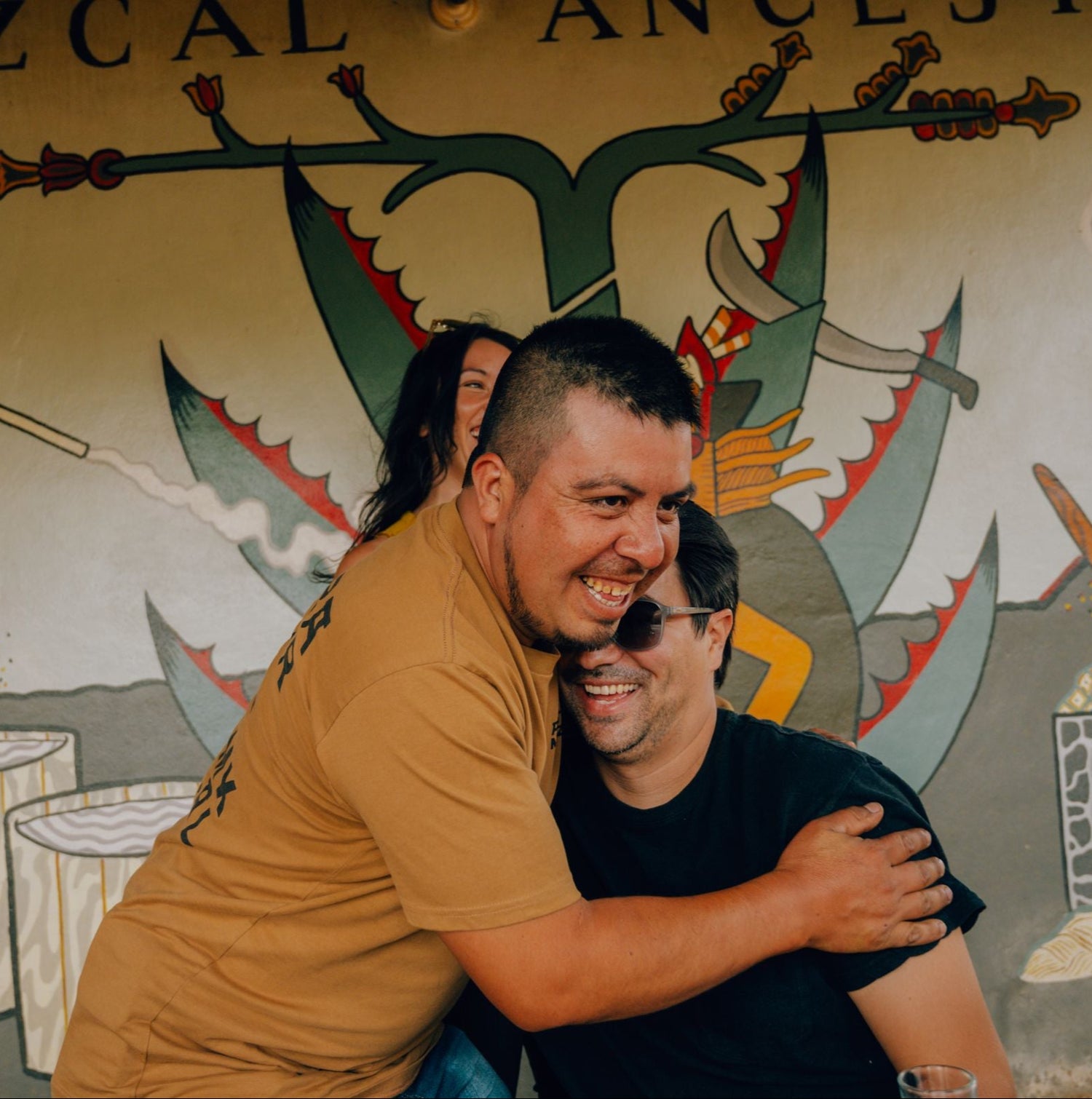 Two men hugging in front of a decorative wall with artistic elements and text.